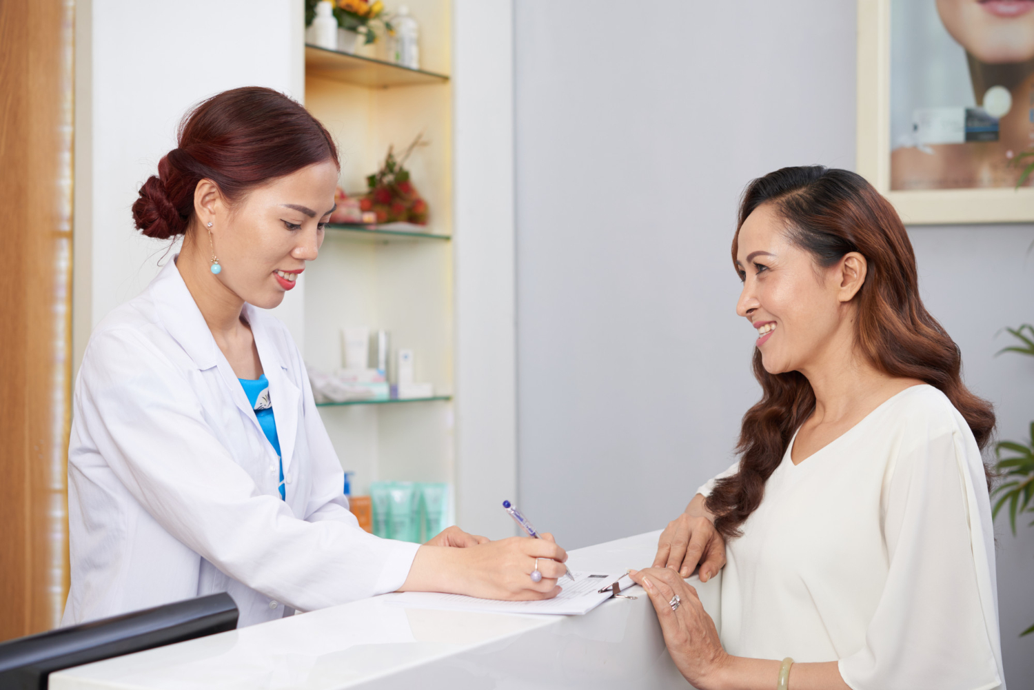 Female healthcare professional in a white coat signs a form for a smiling female patient at a clinic reception surface near a shelf of products.
