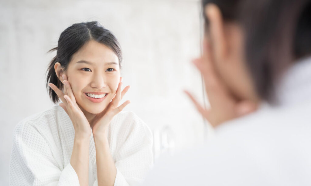 Asian woman in a white robe smiles at her reflection in a mirror, cupping her cheeks with both hands in a bright bathroom setting.