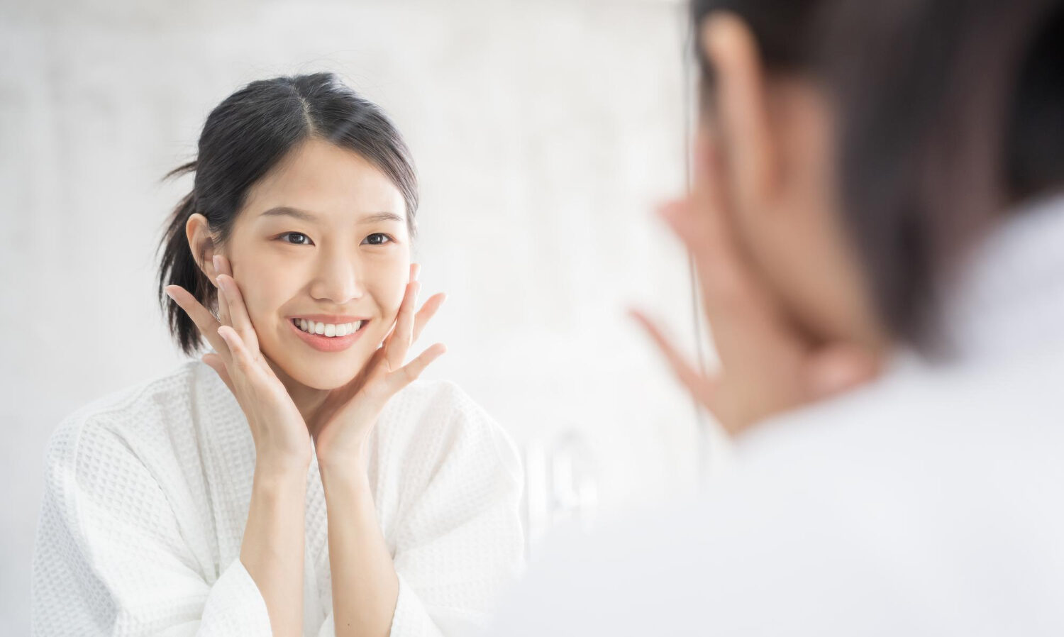 Asian woman in a white robe smiles at her reflection in a mirror, cupping her cheeks with both hands in a bright bathroom setting.