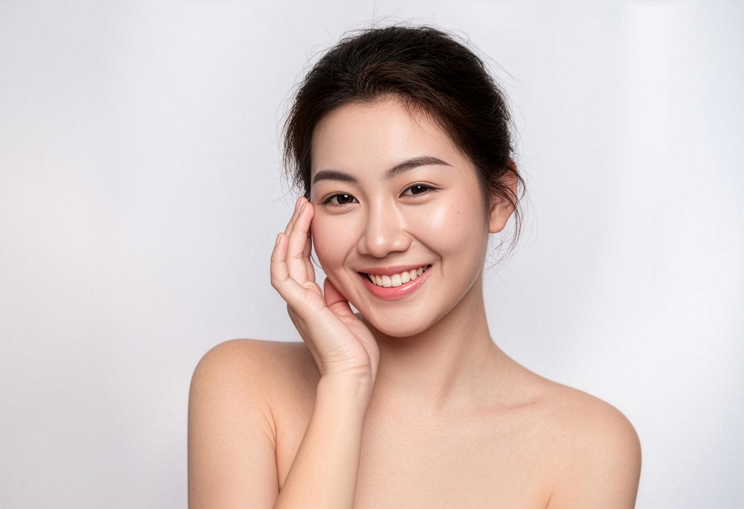 Smiling Asian woman with bare shoulders touching her cheek against a light gray backdrop, portrait photo.