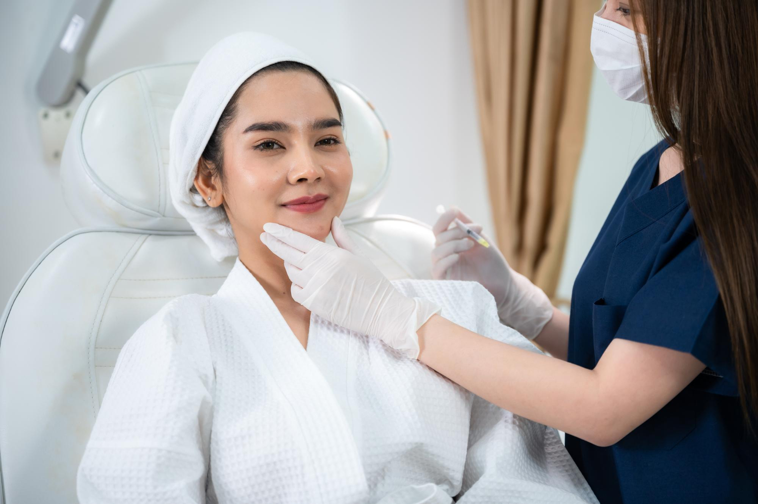 Patient receiving a cosmetic facial injection: clinician in mask and gloves administers treatment near the cheek while patient smiles in a white robe and towel cap.