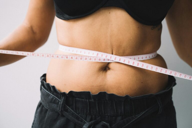 Person's back wearing a black top with a measuring tape wrapped around the waist, measuring around the midsection for waist size.